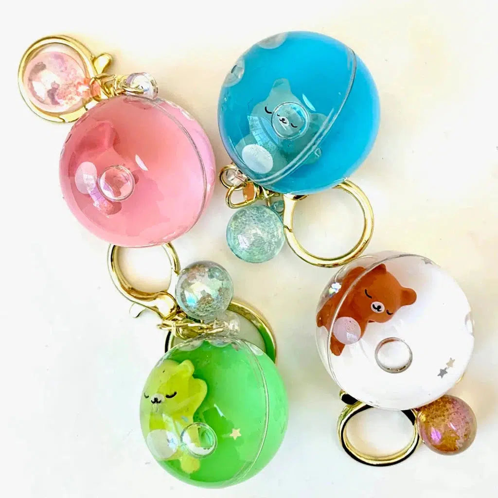 Four colorful spherical keychains with small bear figures inside: pink, blue, green, and white. Each has a gold keyring and decorative bubbles.