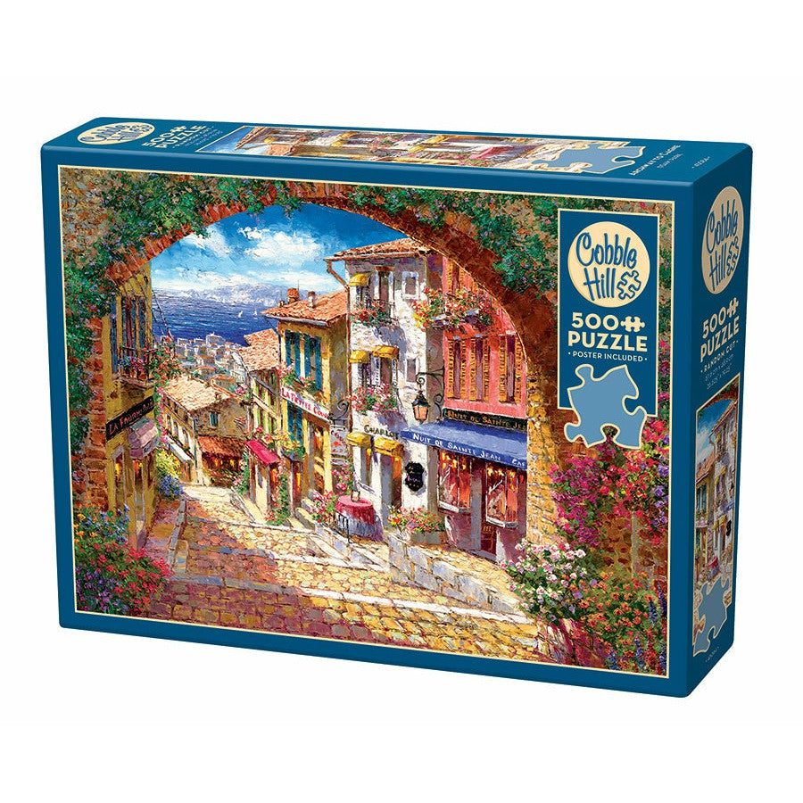 Box of a 500-piece Cobble Hill puzzle featuring a colorful, quaint street scene with a floral archway, balconies, shops, and a distant view of the water.