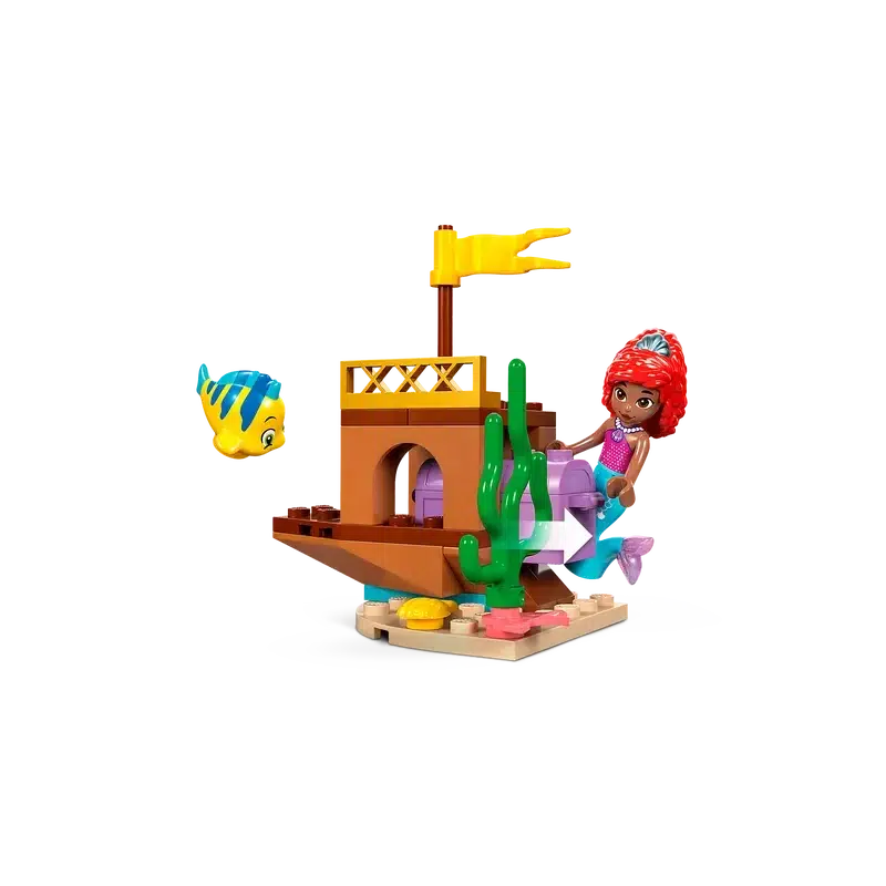 In this enchanting toy scene, a red-haired mermaid reminiscent of Disney characters lounges beside a small brown building toy with a yellow flag on top and green plant elements. Nearby, a fish with blue stripes weaves through the vibrant display, capturing the magic of LEGO creativity.