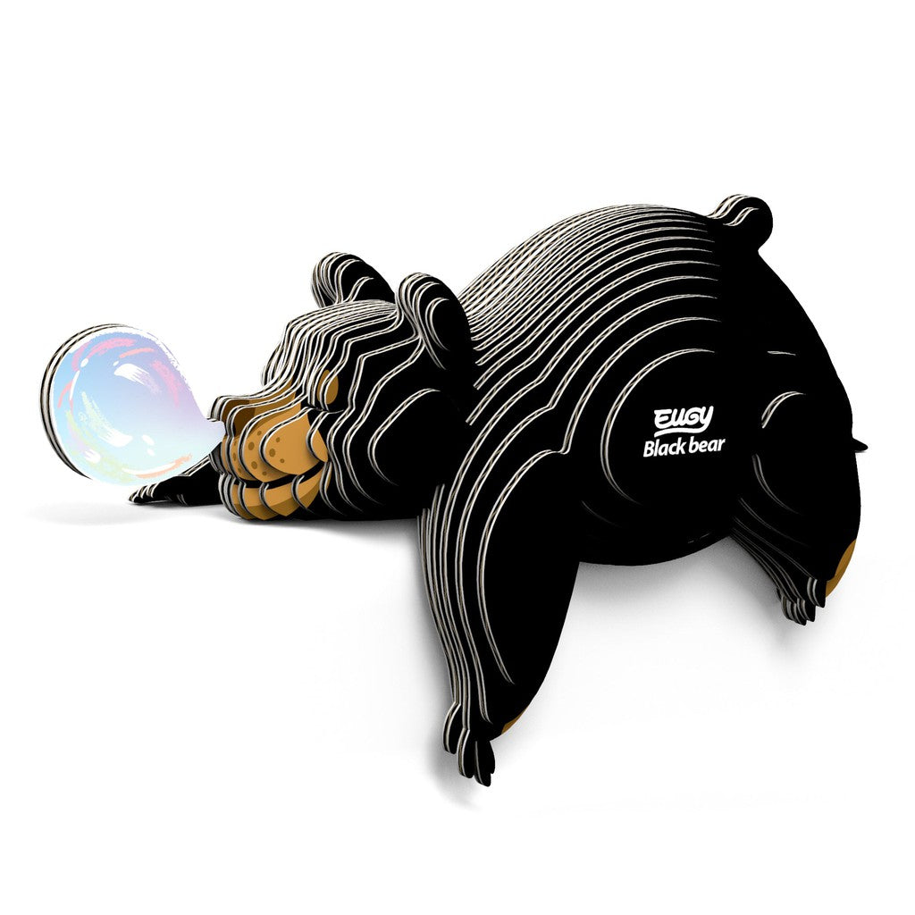 Illustration of a black bear, crafted like an eco-friendly collectable toy, made of layered, striped materials, lying down and blowing a translucent bubble.