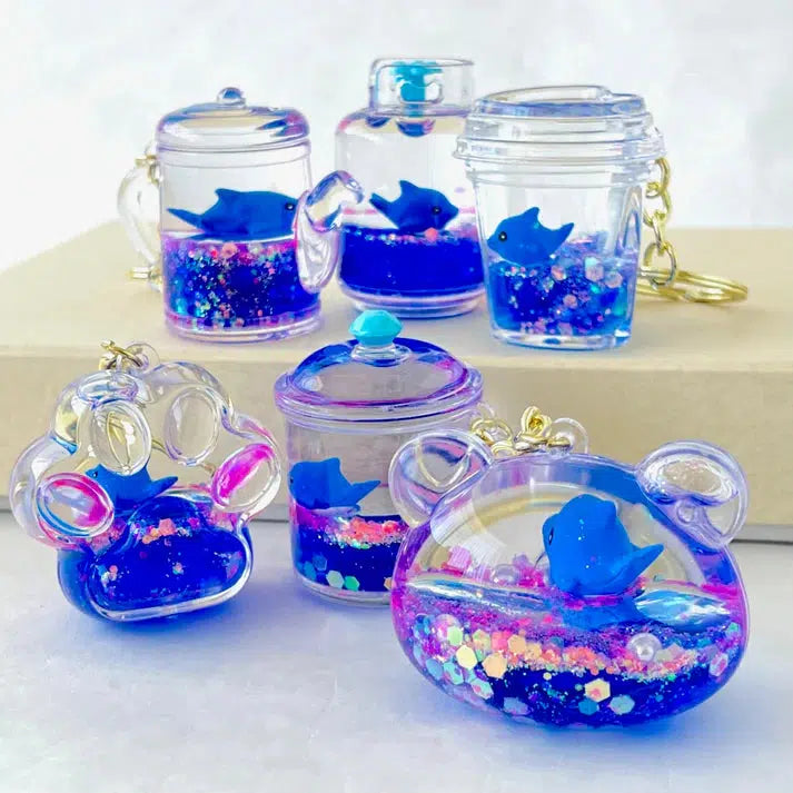 These colorful resin key charms boast assorted designs, featuring mini blue dolphins and silly animal shapes. Resembling drinks and paw shapes, they are filled with blue liquid and glitter for a whimsical touch.