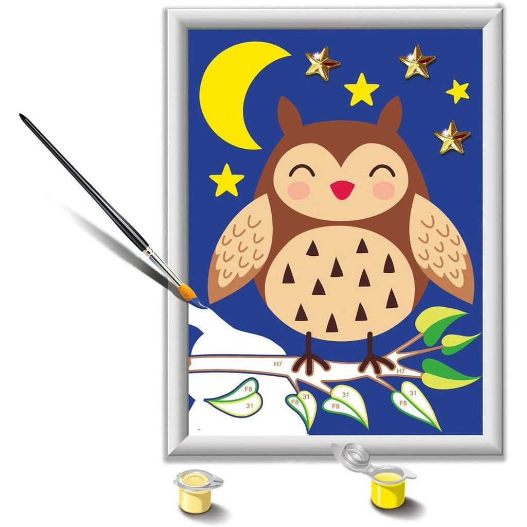 A painting of a cartoon owl on a branch under a moon and stars, accompanied by a Ravensburger CreArt kit with paintbrush and paint pots, invites creative minds to explore their own artistic adventures.