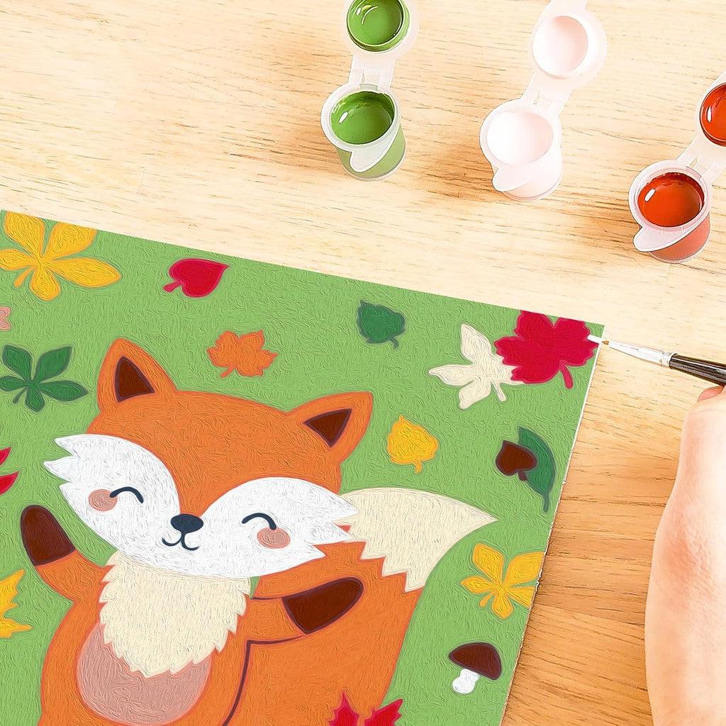 A person engages in arts and crafts, painting a colorful autumn-themed canvas of a cute fox surrounded by leaves. Open paint pots in green, white, and orange evoke the charm of a classic Ravensburger CreArt set.