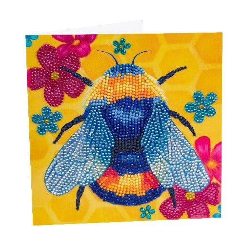 A card with an image of a bee. The bee is decorated with crystals from the kit and is surrounded by flowers