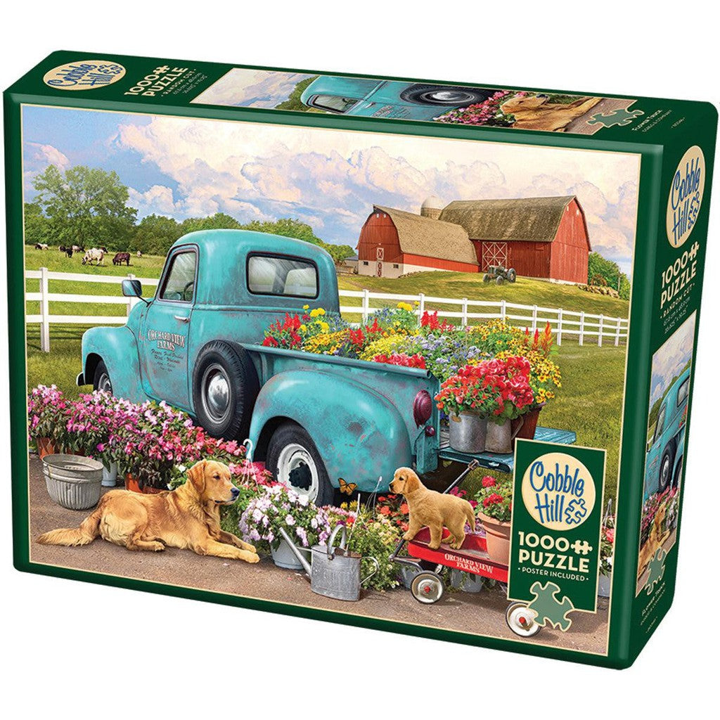 1000 pc puzzle of a blue old-fashioned truck parked near a barn. The truck bed is bursting with colorful flowers and there are two golden Labradors near the truck