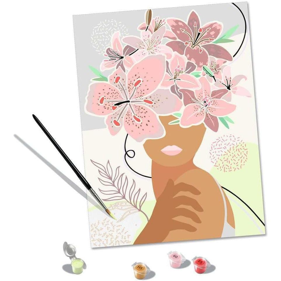 A painting titled "Flowers on My Mind" features a woman with large pink blossoms instead of hair. A Ravensburger Paint by Numbers kit, including a paintbrush and small pots of paint, sits beside the canvas, inviting creativity and artistry.