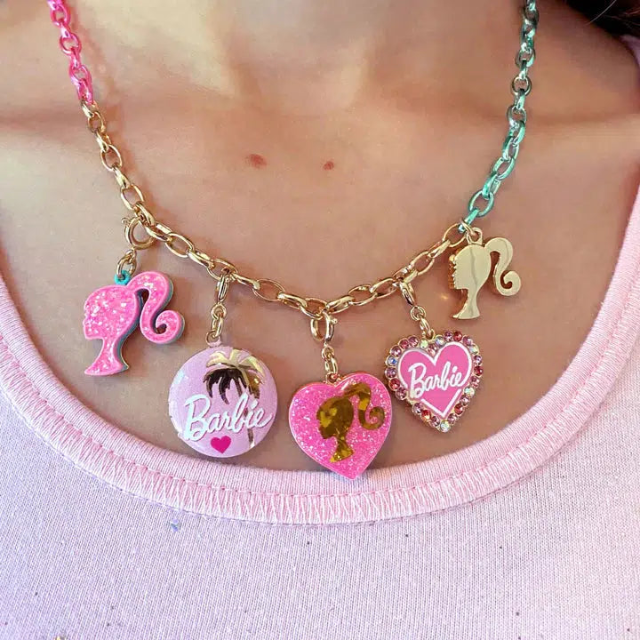 A person wearing a pink top and a gold chain necklace with Barbie-themed charms from CHARM IT!, including silhouettes and hearts, some adorned with glitter and rhinestones, perfect for Barbie fans.