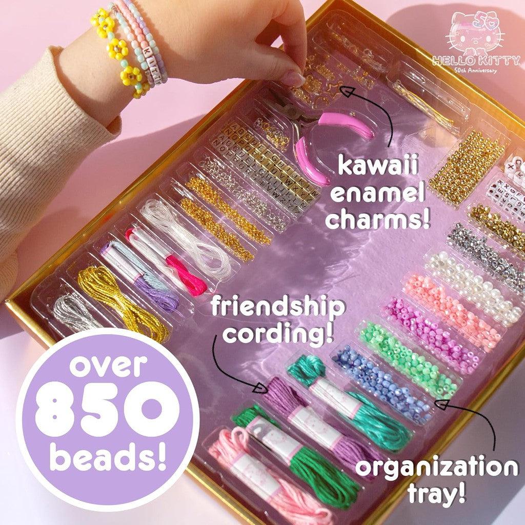 A person's hand wearing bracelets hovers over a Jewelry Making Kit filled with colorful beads, kawaii enamel charms, and friendship cording in an organization tray. Text highlights features and bead count alongside adorable Hello Kitty jewelry components.