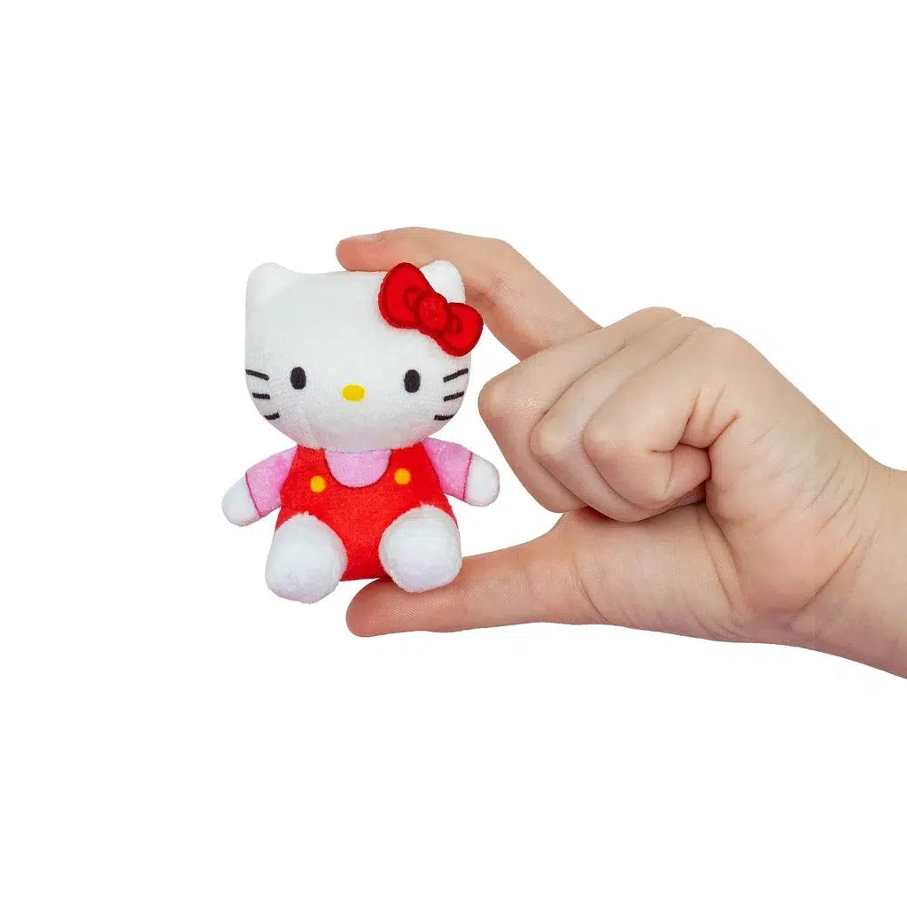 A hand holds a small plush toy of a white cat that resembles Hello Kitty, adorned with a red bow and wearing a red outfit.