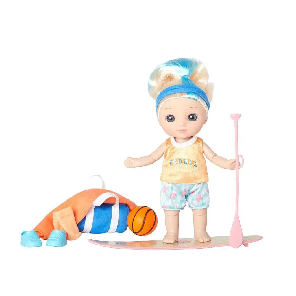 A blonde doll in a ponytail, sporting a headband, orange top, and shorts, stands confidently on her paddleboard with a pink paddle. Nearby, blue shoes rest beside a rolled towel and an orange-striped basketball.