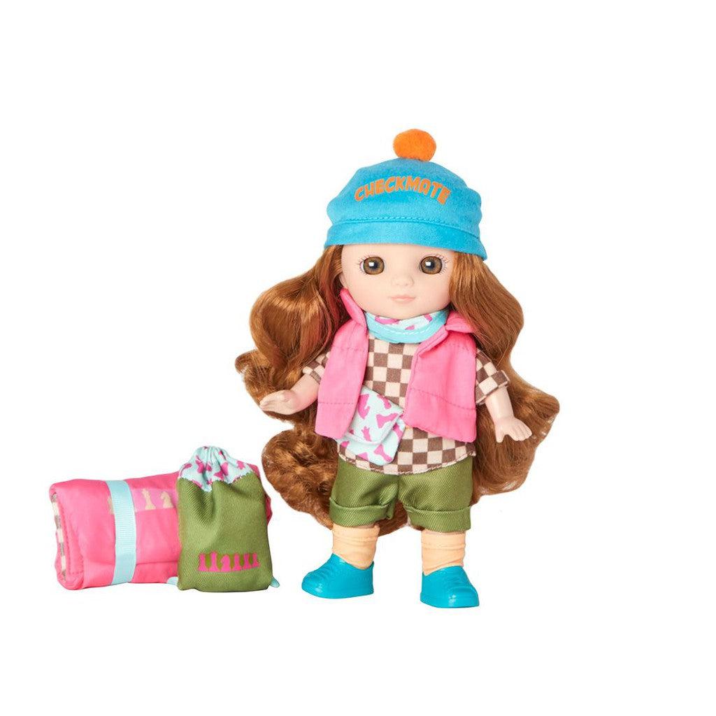 Doll with long brown hair, wearing a pink vest, green shorts, blue shoes, and a blue hat. Beside it is a rolled-up pink and green sleeping bag, perfect for camping adventures.