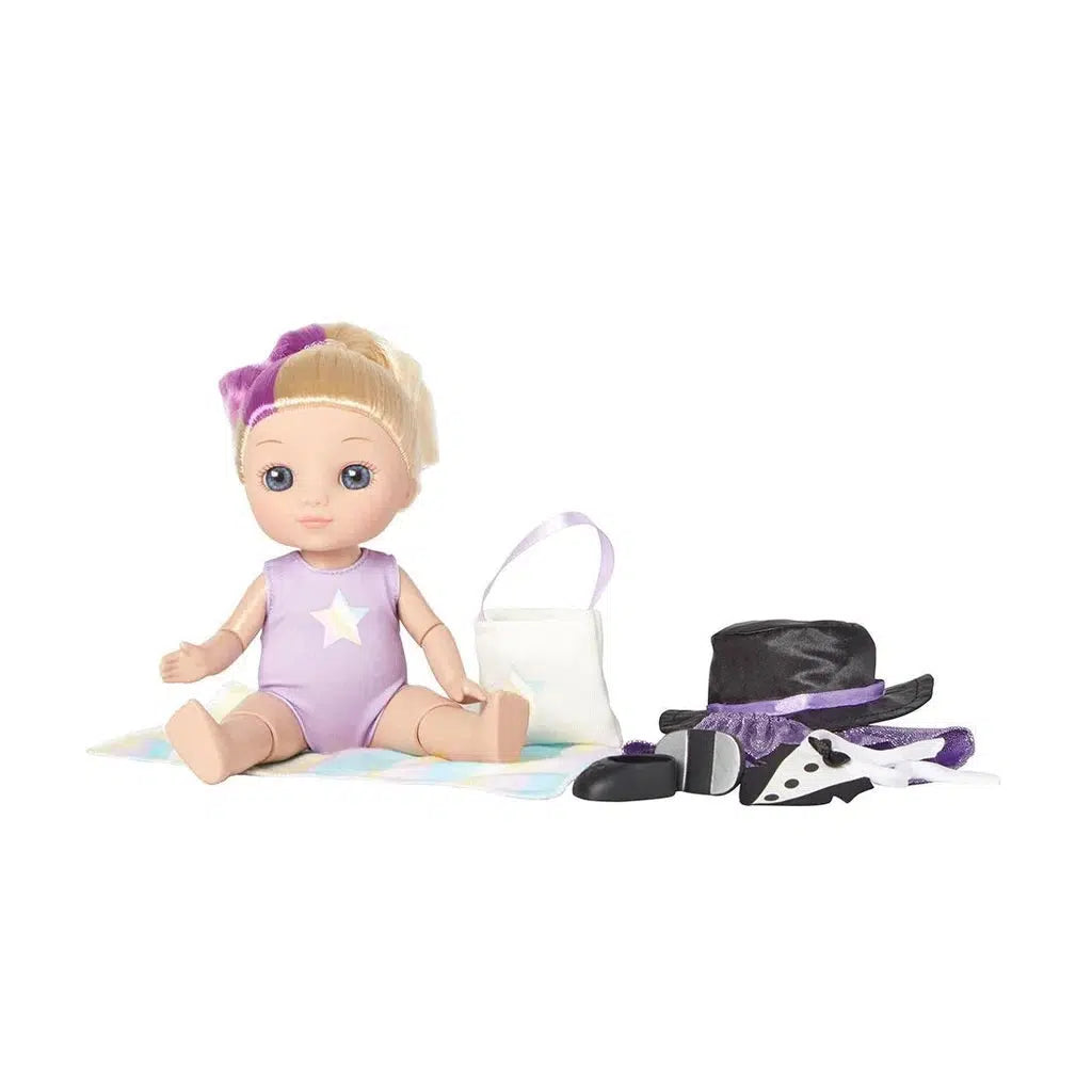 A doll wearing a purple outfit sits gracefully on a patterned mat, surrounded by accessories for her next performance: a hat, mask, and a small bag for the dance recital.