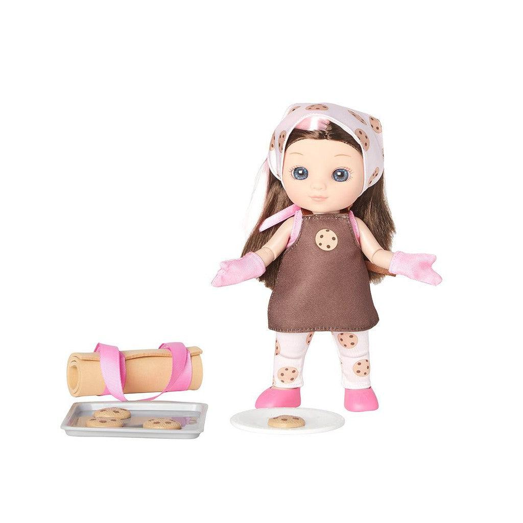 A posable baker doll wearing a brown apron and cookie-patterned headscarf stands next to a tray, cookies, a rolling pin, and a plate with a cookie on it.