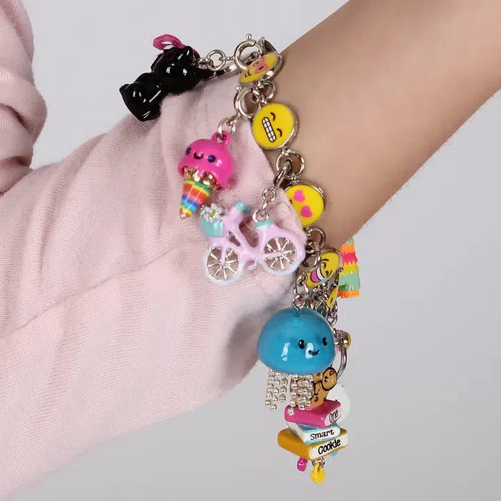 Close-up of an arm wearing a charm bracelet adorned with playful designs, including a pink ice cream, a blue cloud, and a delightful jellyfish charm. Perfect for the ocean animal lover, other charms feature yellow emojis and a black cat on the chain.