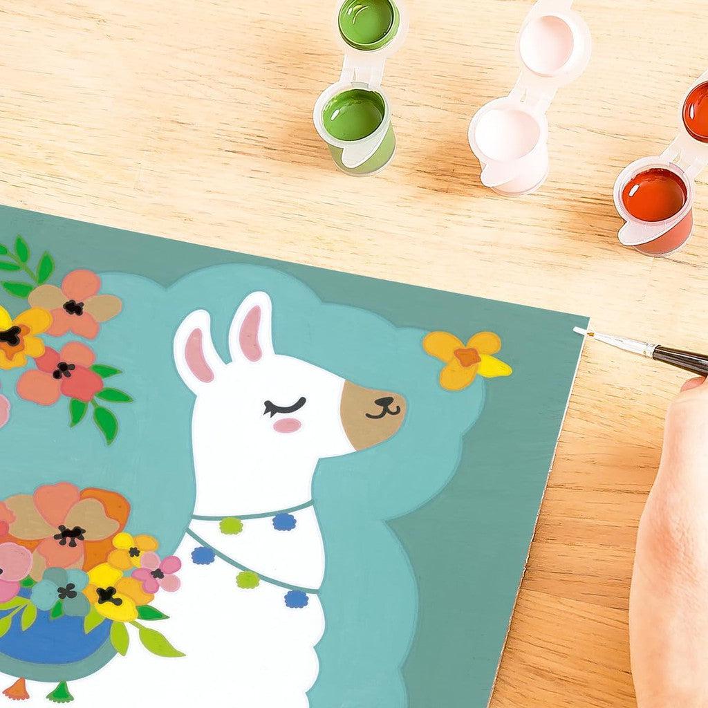 A person diligently painting a colorful llama illustration with a small brush, reminiscent of a Paint by Numbers kit. Nearby, paint pots filled with green, white, and orange rest on the wooden surface—a scene that embodies the joy of arts and crafts.