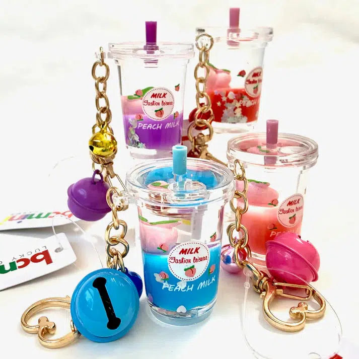 Four colorful milkshake keychains with bells and charms are displayed. Each features a different color lid and liquid inside, labeled "Peach Milk." With assorted designs, these whimsical Peach Milk floaty key charms add a playful touch to any collection.