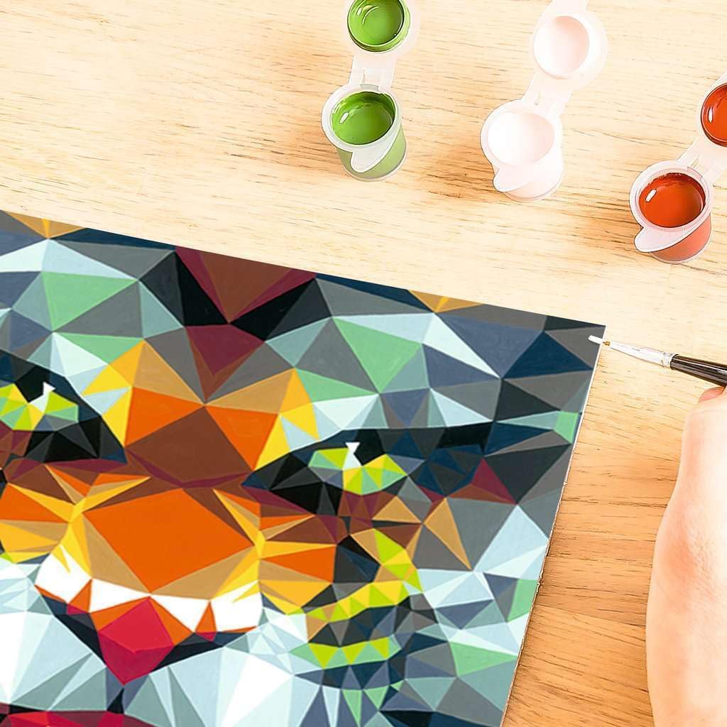 A partially completed geometric painting of a lion's face, reminiscent of a Ravensburger Polygon Tiger, rests on a wooden table, surrounded by three small open paint pots in green, brown, and red.