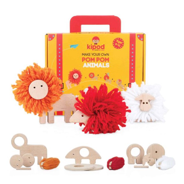 Wooden toy set with pom-poms shaped like animals, including a lion and a sheep. The box in the background features product branding and the text "Make Your Own Pom-Pom Animals." 