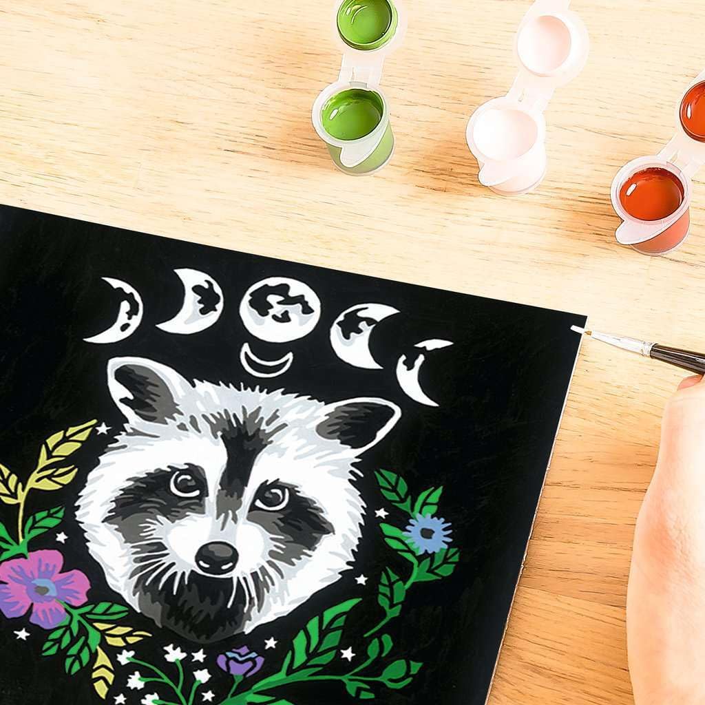 Hand painting an image of a raccoon with a floral wreath and moon phases on a black canvas, using the Raccoon Paint Kit. Paint pots and a brush rest on the wooden table, making it easy to dive into this creative journey.