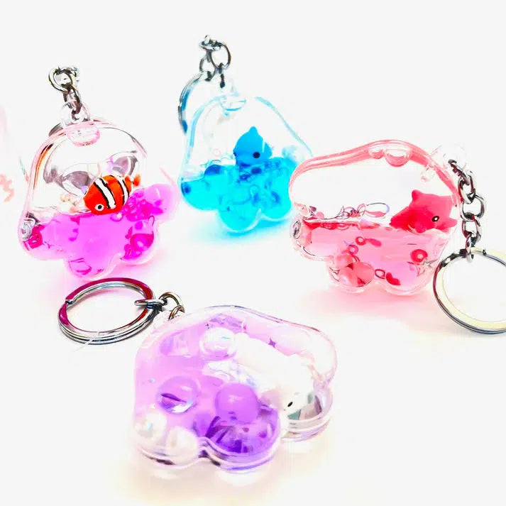 Four transparent FLOATY KEY CHARMS in pink, blue, and purple, each holding small animal figures and liquid inside. Two keychains feature fish and two showcase silly penguins, all surrounded by tiny bubbles for a playful touch.