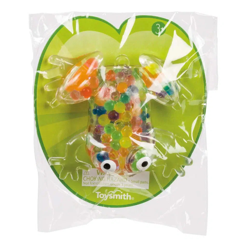 A clear plastic frog squish toy brimming with colorful beads offers a delightful gooey squeeze effect, packaged in a sealed bag. The packaging features safety warnings and proudly bears the "Toysmith" label.