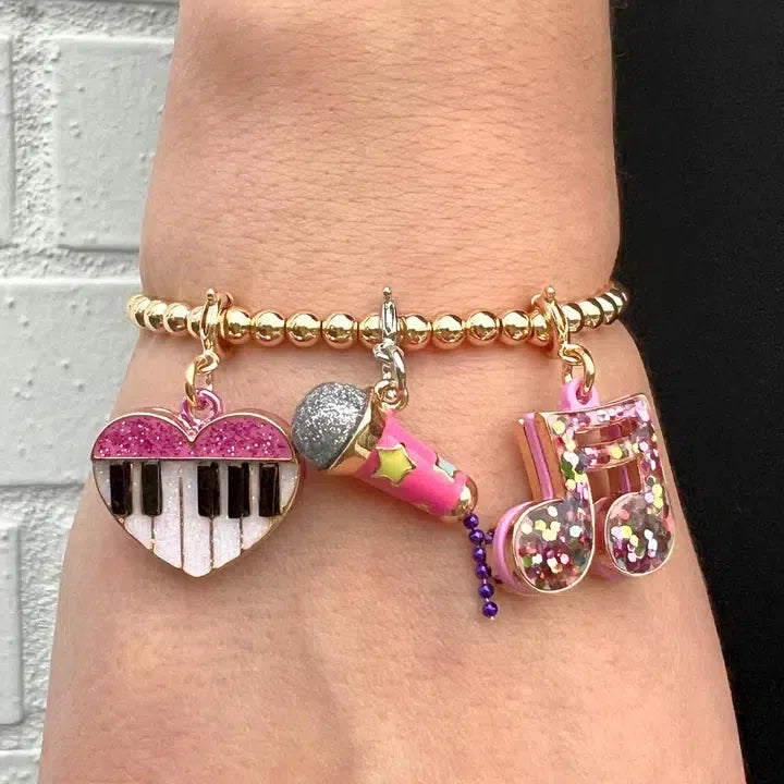 This bracelet, perfect for a music lover, features gold beads and three enchanting charms: a heart-shaped piano key, a pink microphone charm, and musical notes adorned with multicolored sparkles.