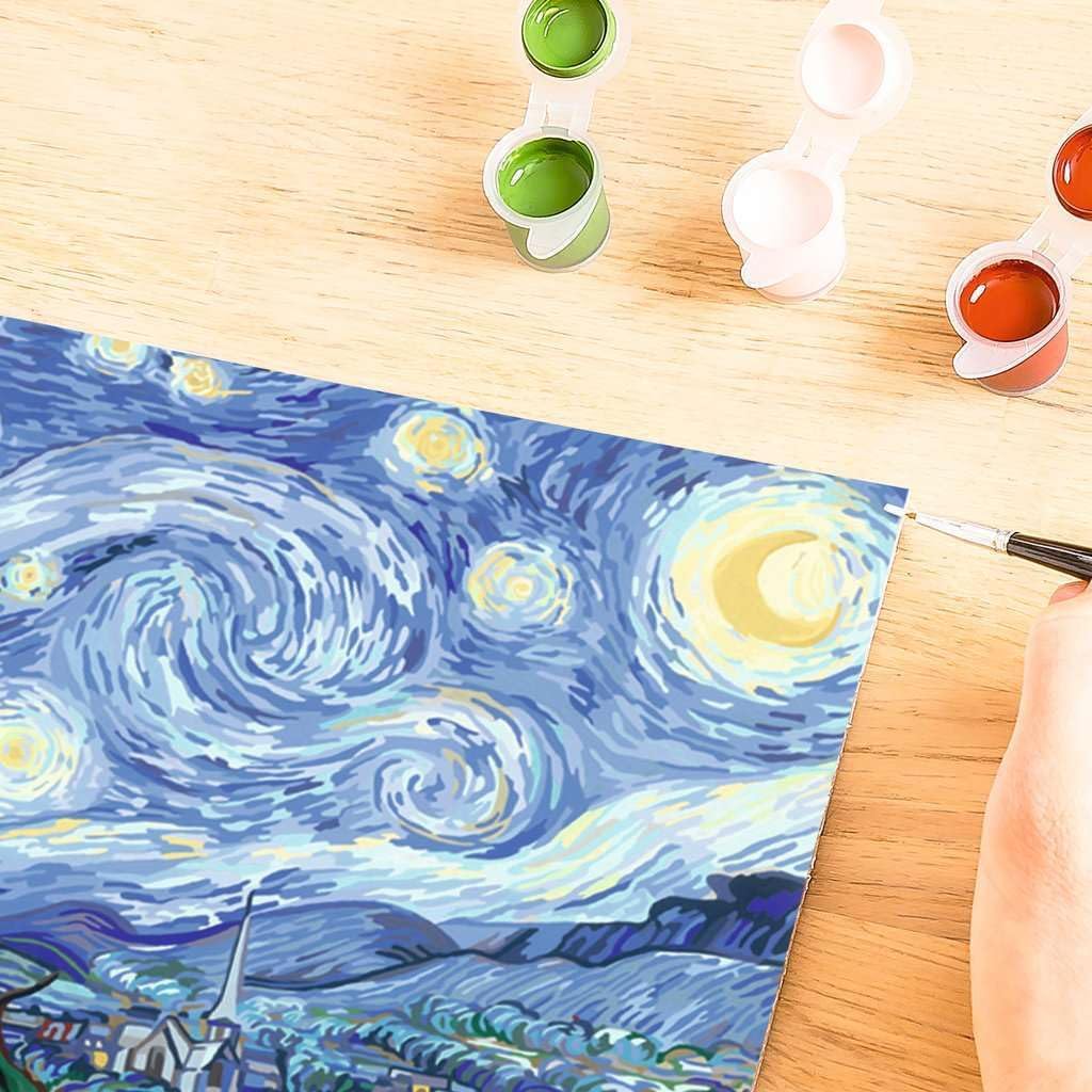 A person delicately uses a paintbrush to recreate "The Starry Night" on canvas, reminiscent of a Paint by Numbers Kit. Small open paint pots in green and red are scattered across a wooden table, capturing the magic of a Ravensburger Van Gogh masterpiece.