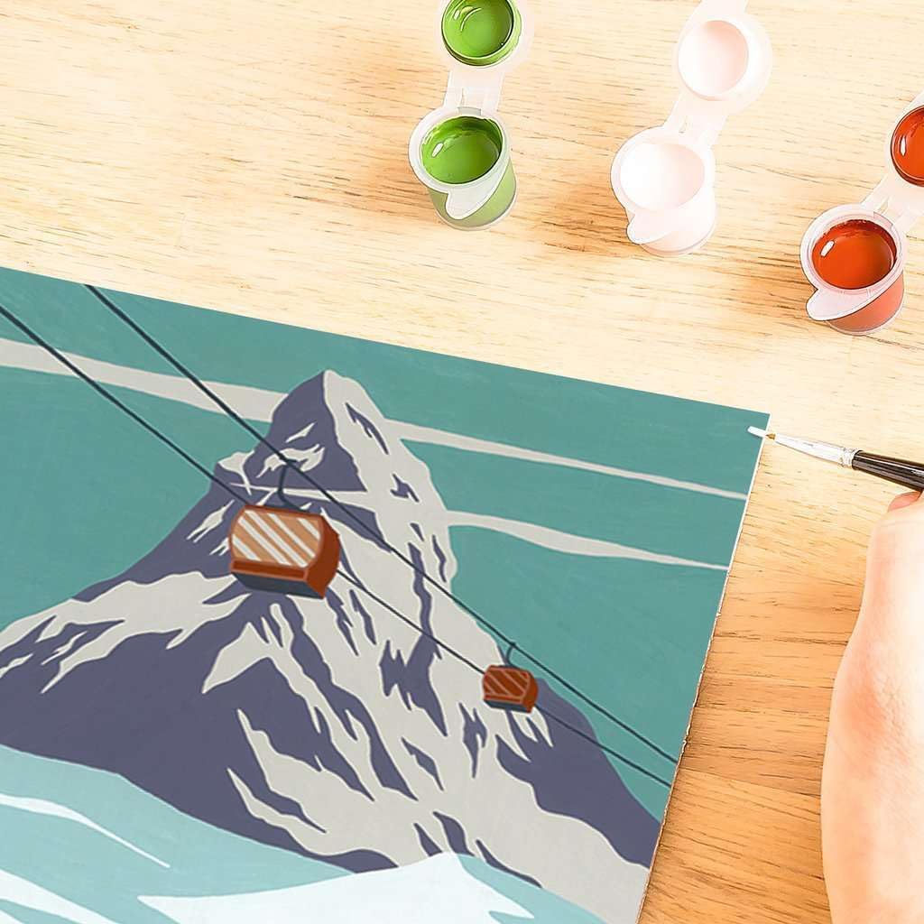 Close-up of a Ravensburger Zermatt paint-by-numbers canvas depicting a snowy mountain with cable cars. A hand delicately holds a brush, while small paint pots sit invitingly on the wooden surface, epitomizing the joy of Painting Arts and Crafts.